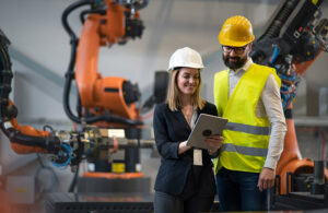 women in a hardhat holding an ipad in front of a robot arm.